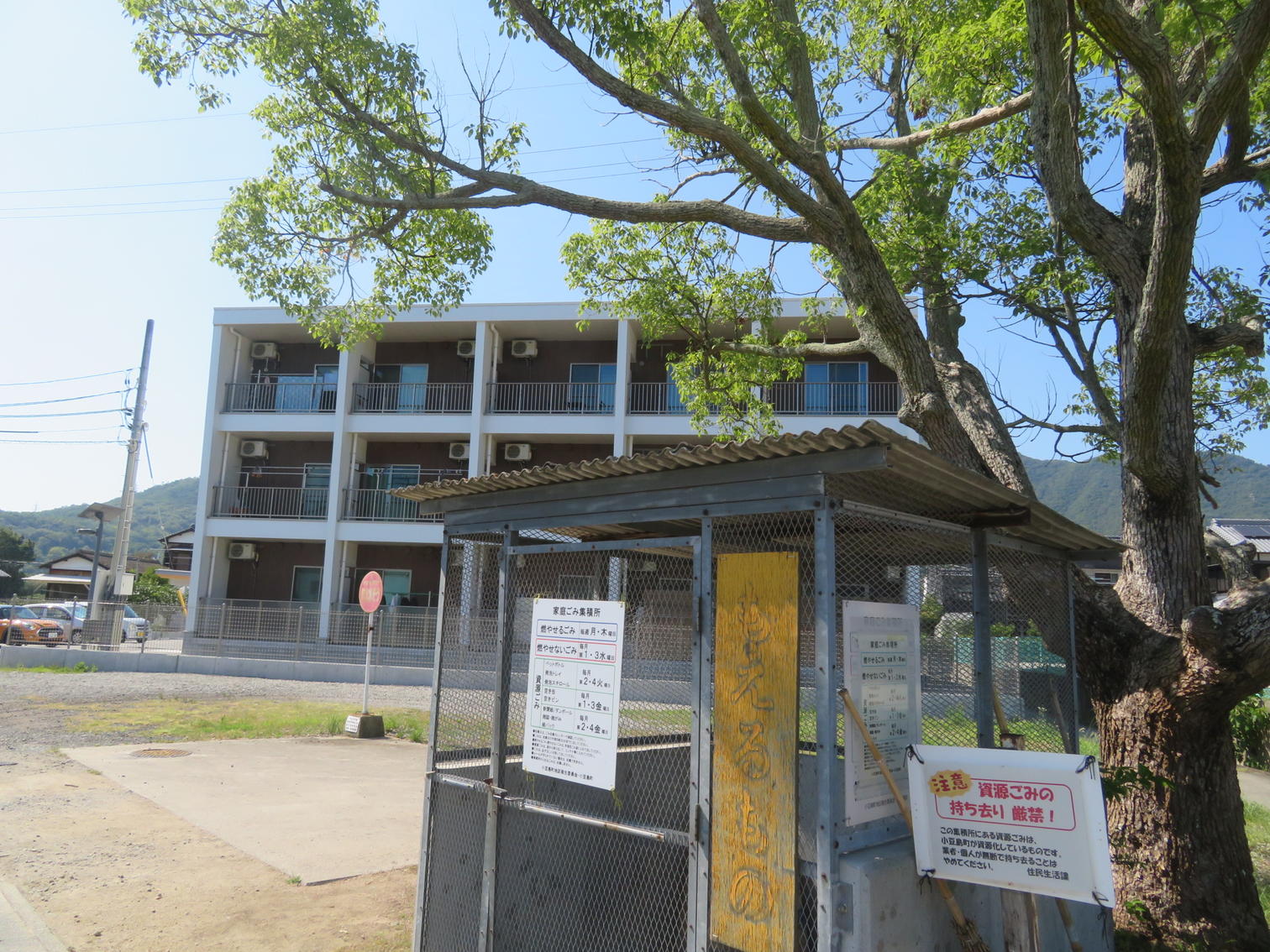 大きな木の下に掲示板があり、背後に集合住宅が見える。